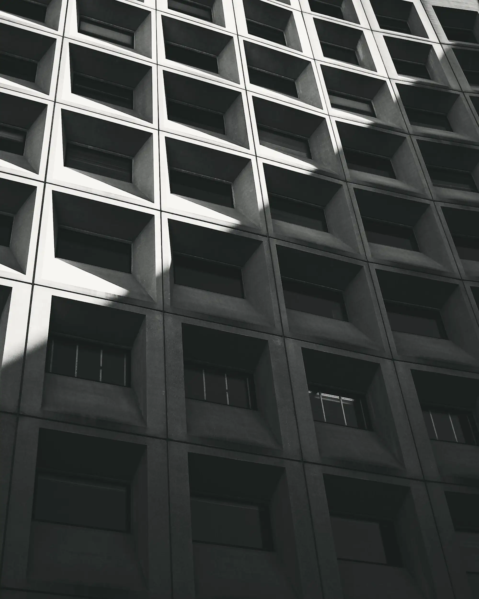 Facade of square cells shot from below, cut diagonally by a band of sunlight.