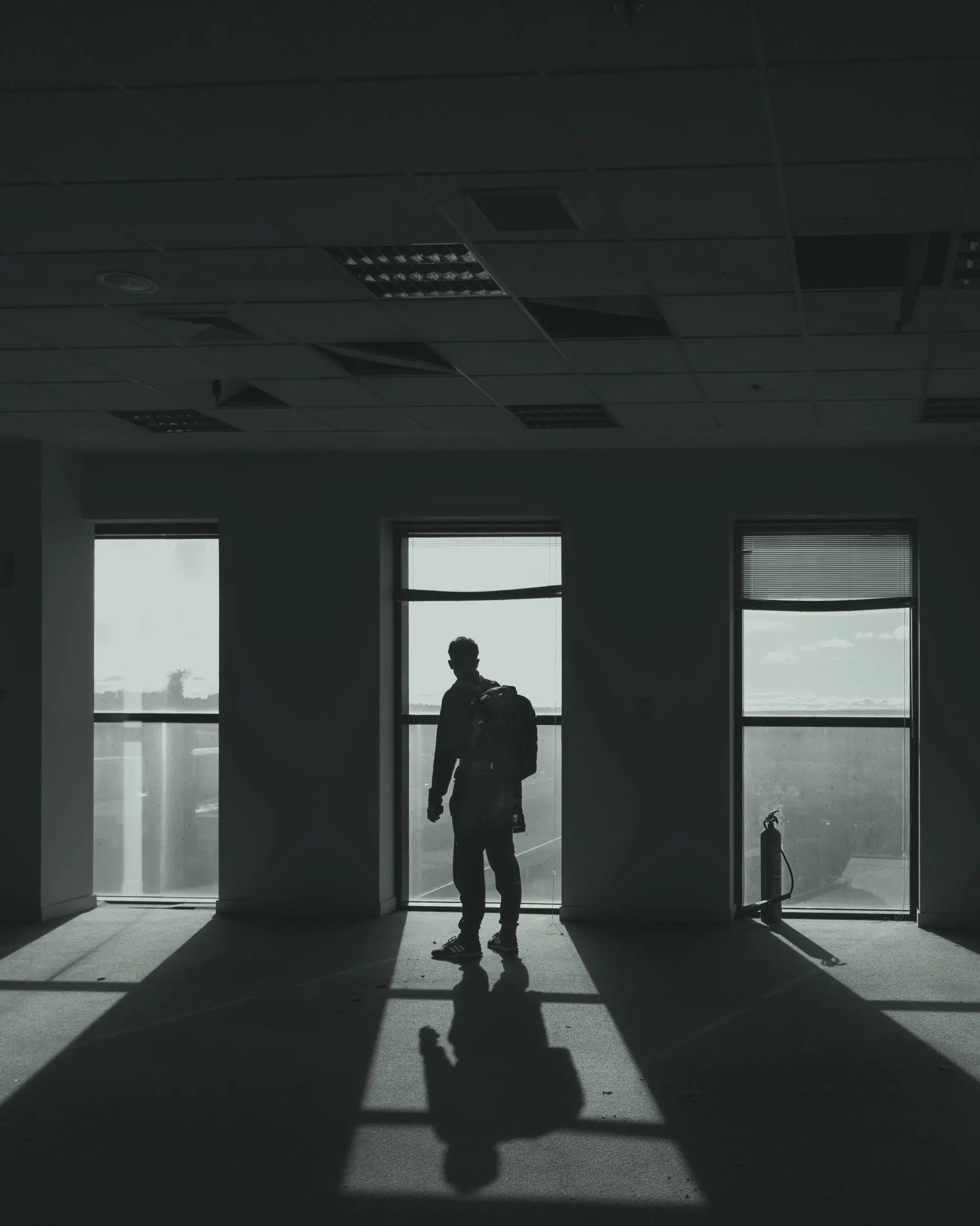 A single figure standing before three tall windows, a long shadow cast across the floor of an empty interior.